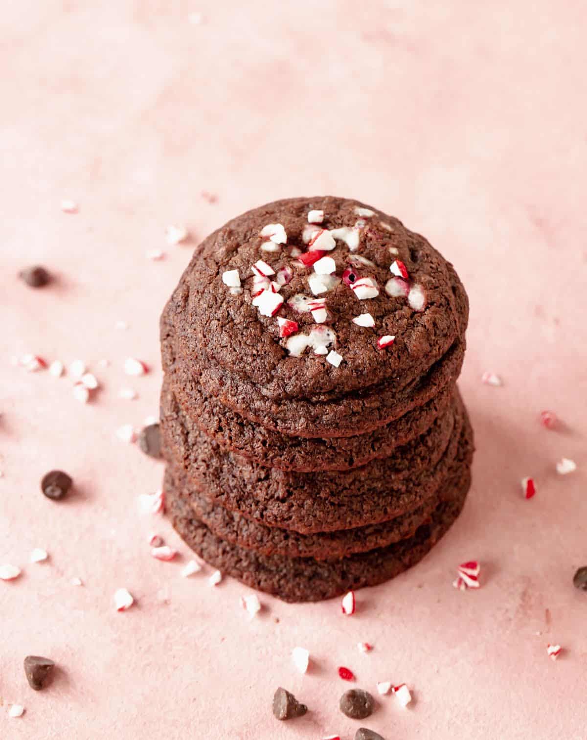 Several chocolate peppermint cookies stacked on pink surface, crushed candy cane and chips around.