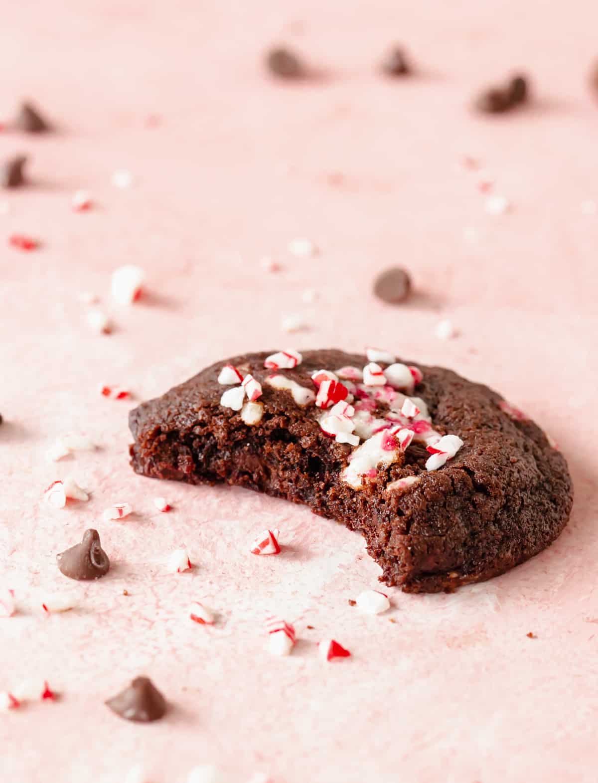 Single bitten peppermint chocolate cookie on pink surface with chips and crushed candy around. 