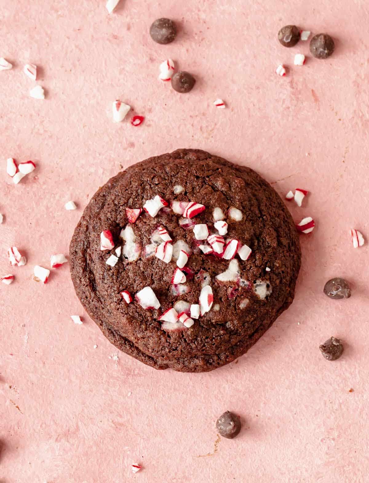 Top view of single chocolate peppermint cookie on pink surface, loose candy and chips around.