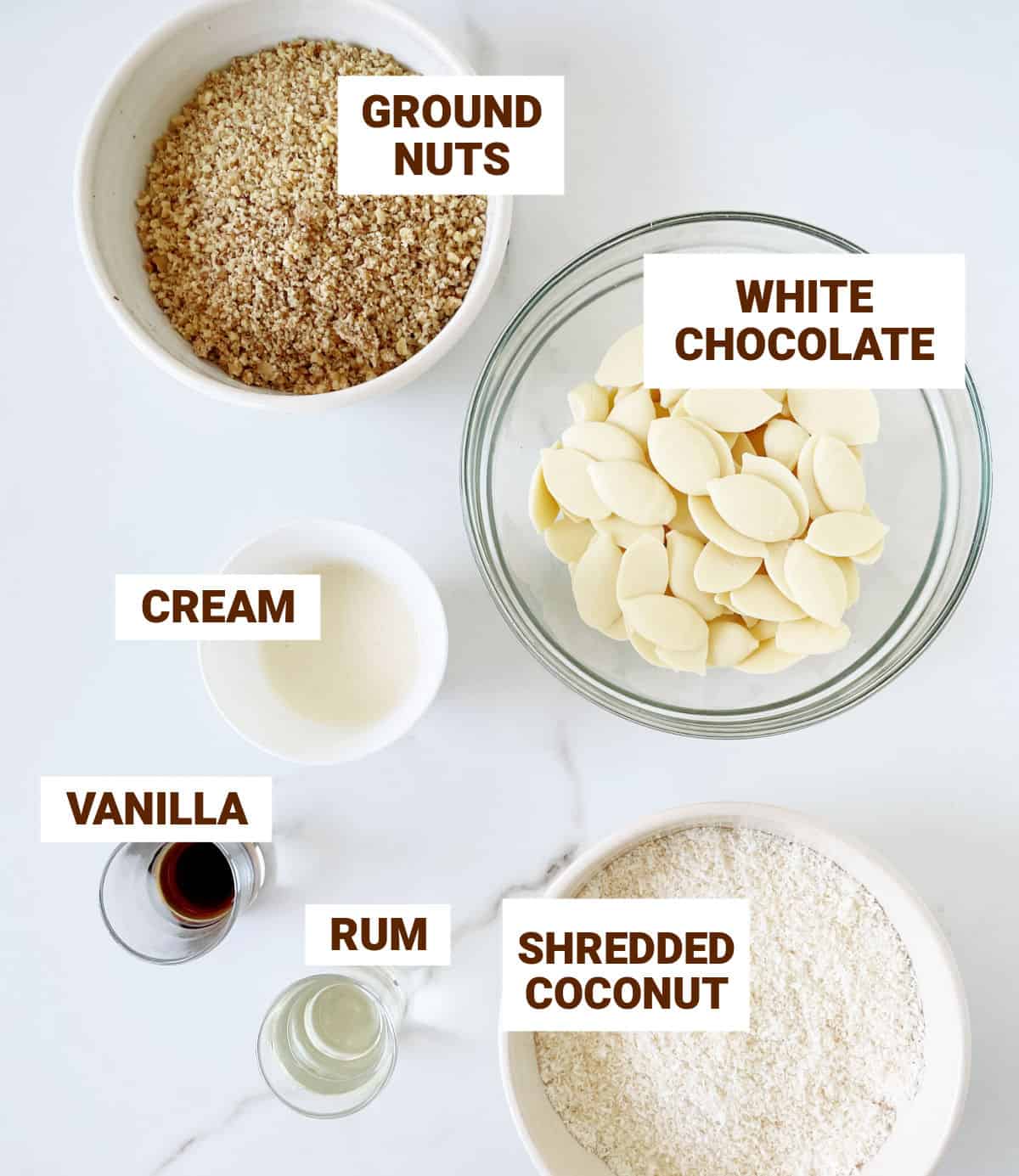 White surface with ingredients in bowls including white chocolate, nuts, coconut, cream, flavorings