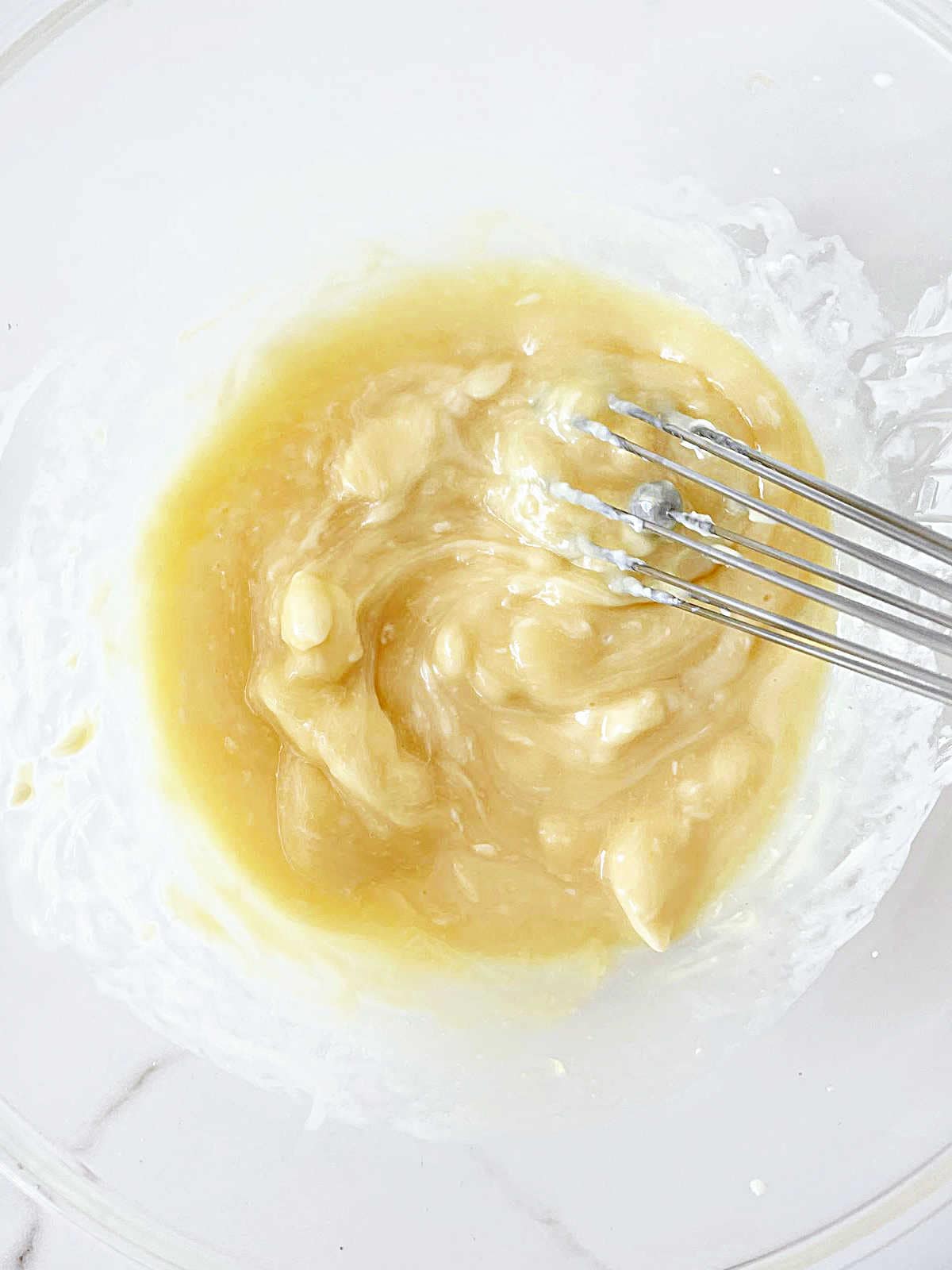 Stirring half melted white chocolate in a glass bowl on a white marbled surface.