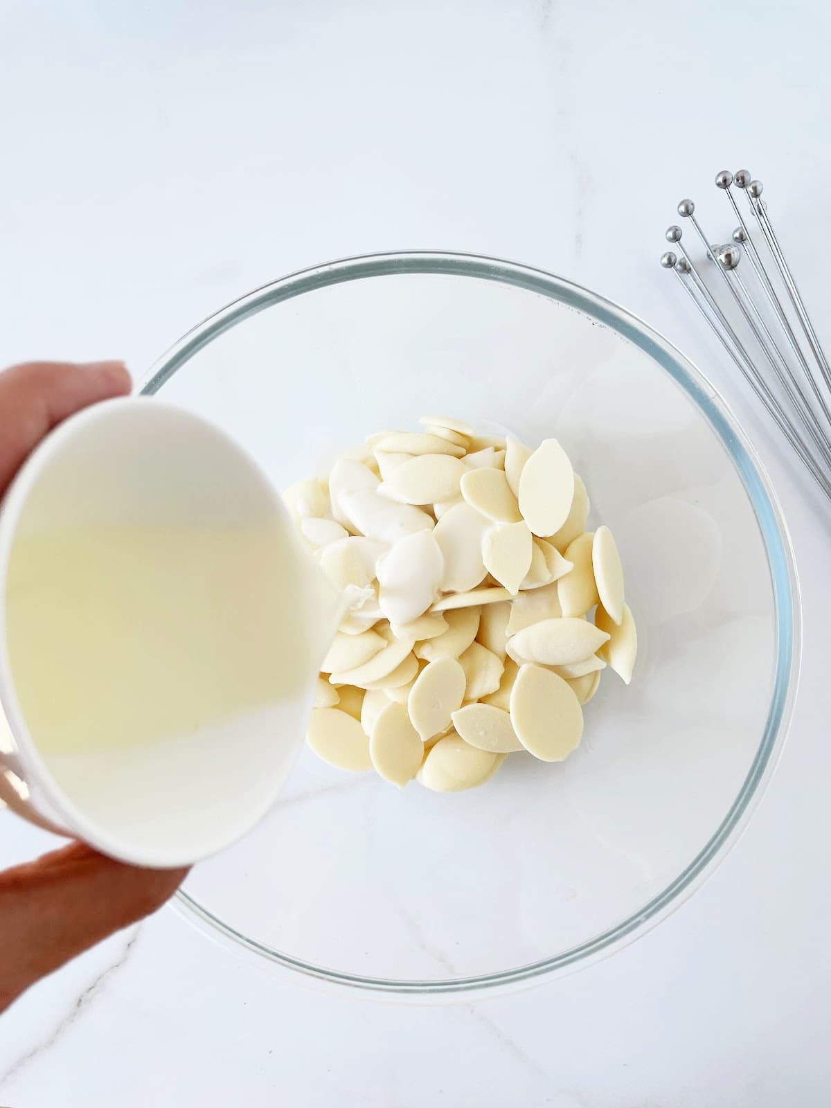 White chocolate pieces in a glass bowl. Cream being poured over. White surface. 