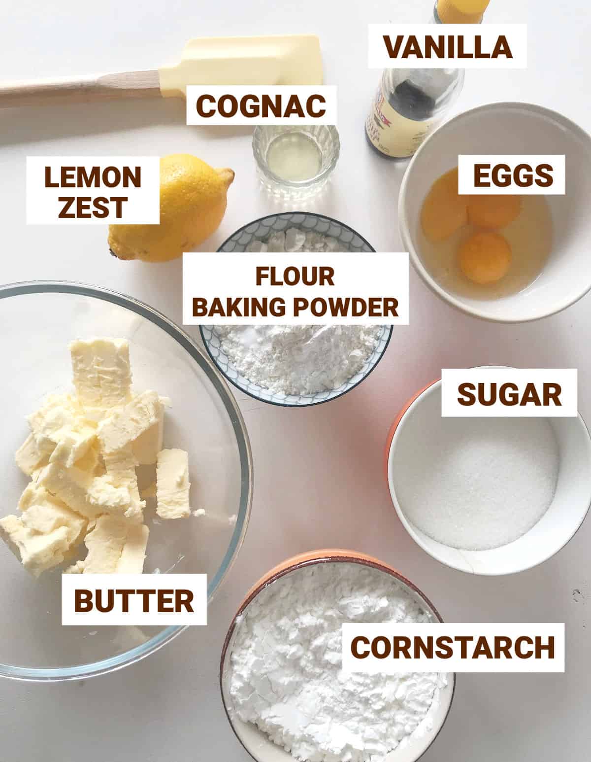 Ingredients for cornstarch alfajores in bowls on white surface, yellow spatula, whole lemon, butter, eggs, sugar, vanilla