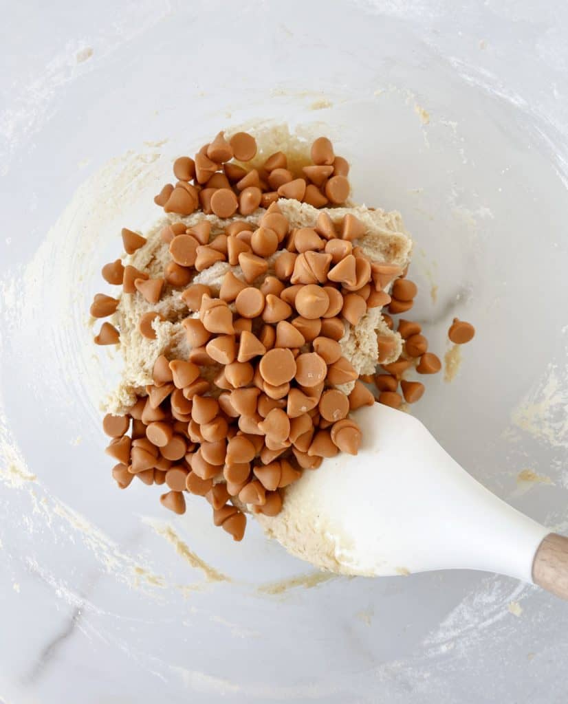 Adding butterscotch morsels with oat cookie dough in a glass bowl. White background.
