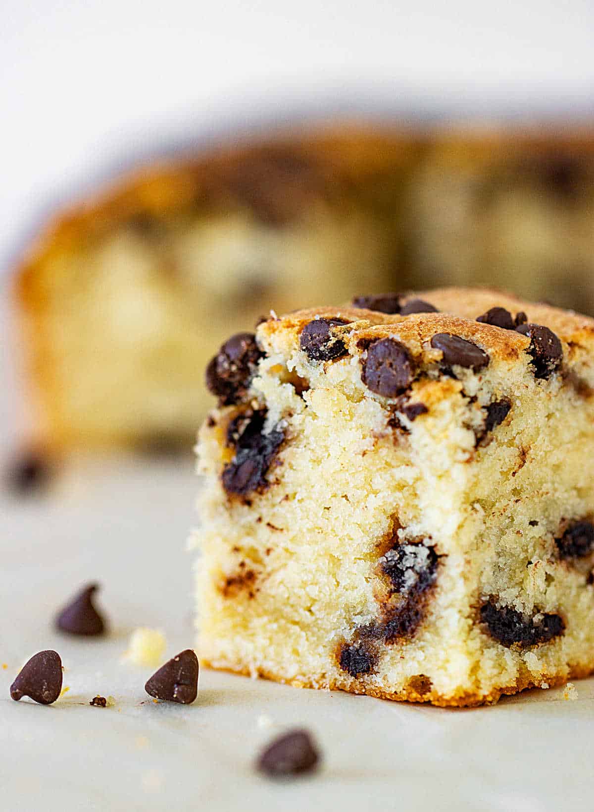 Single square of vanilla cake with chocolate chips on white surface, loose chips around.