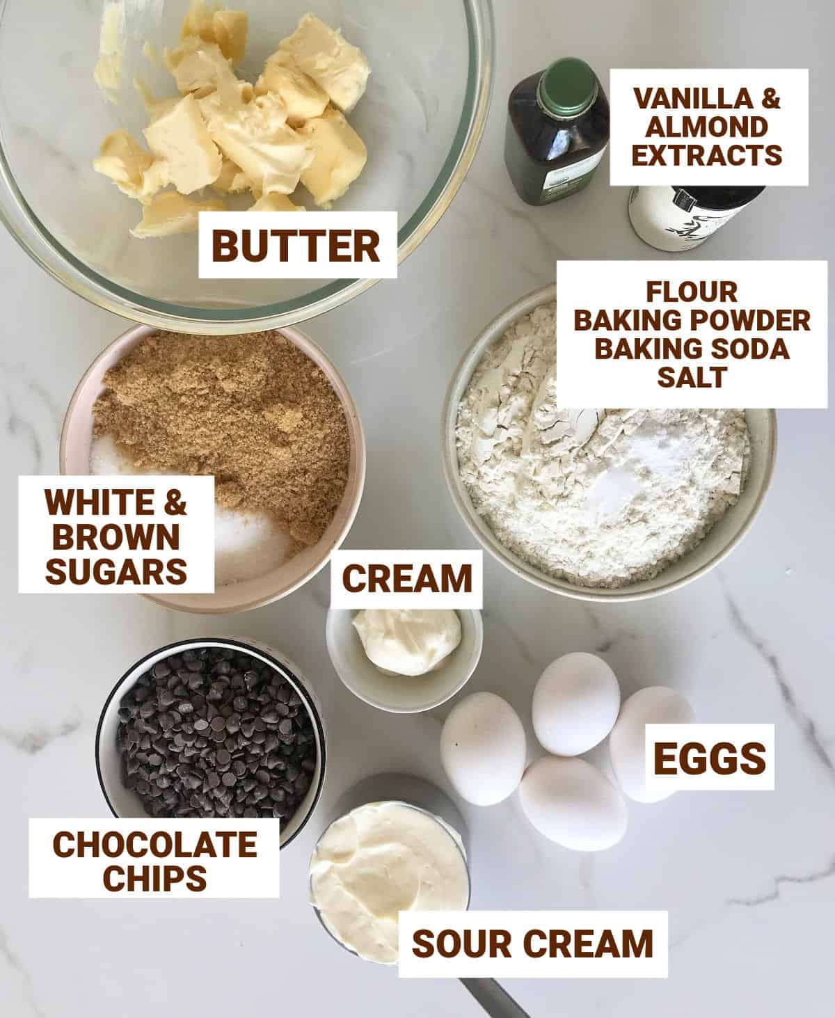 Bowls on white marble surface with ingredients for chocolate chip cake including butter, sugars, flour, eggs, extracts, sour cream.