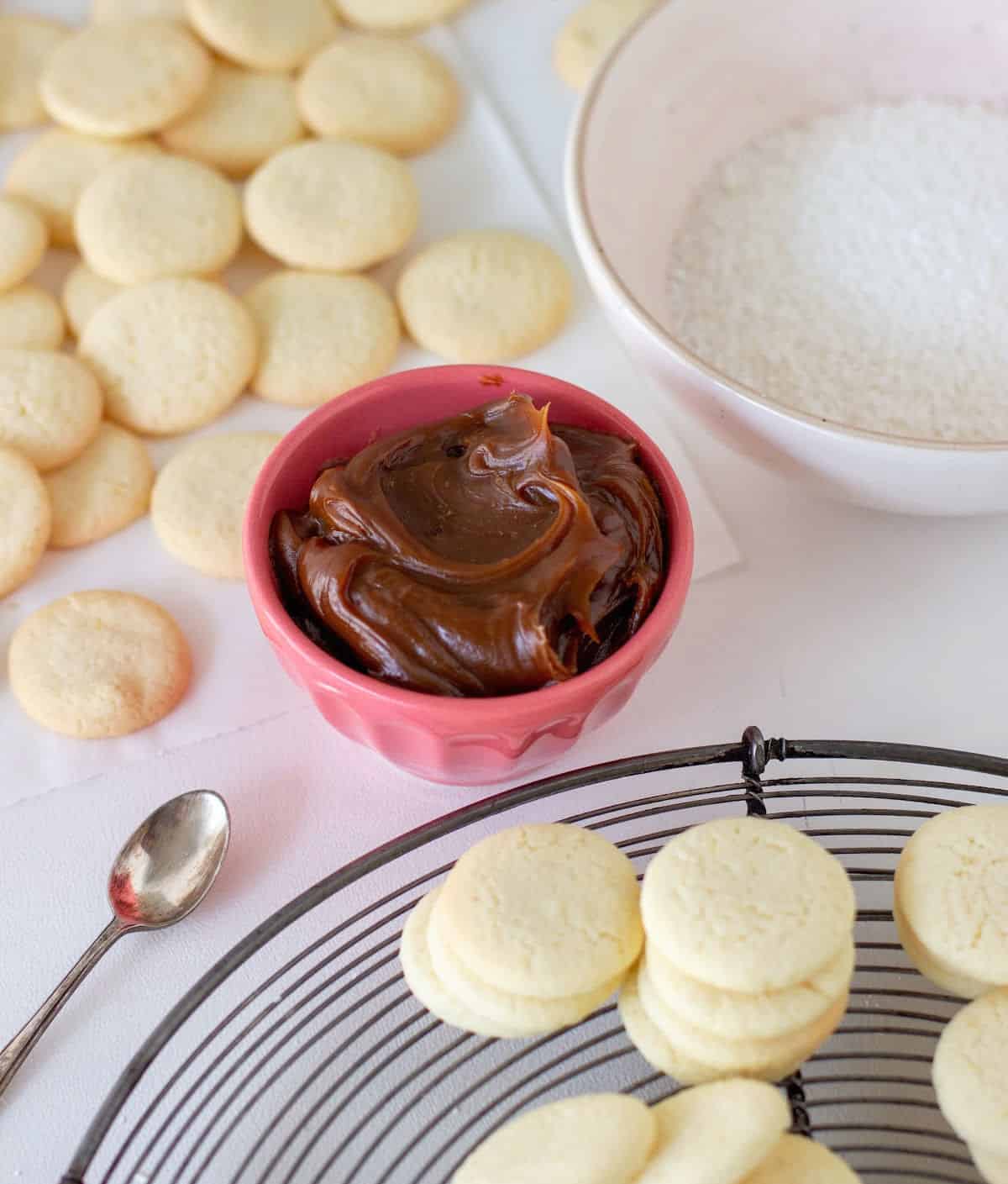 Cornstarch cookies on wire rack, dulce de leche in pink bowl, coconut in bowl, white surface