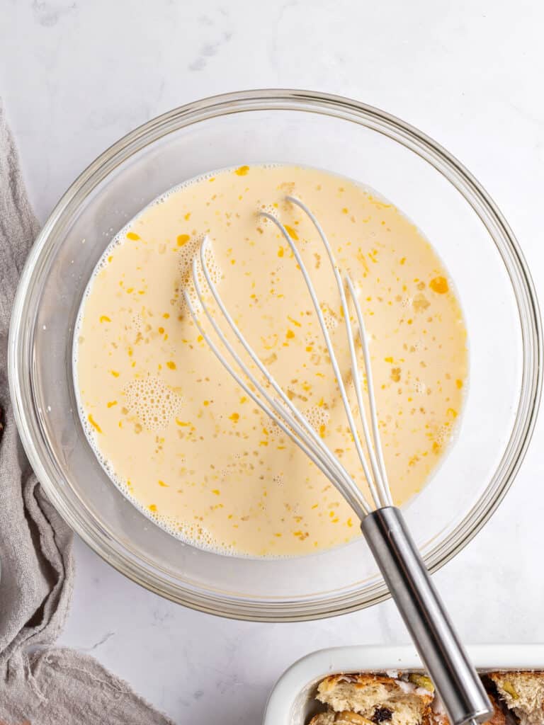 Glass bowl with custard mixture for bread pudding. Wire whisk. Light gray background. Top view.