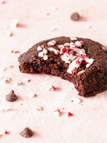 Bitten chocolate peppermint cookie on a pink surface with chips and crushed candy around.