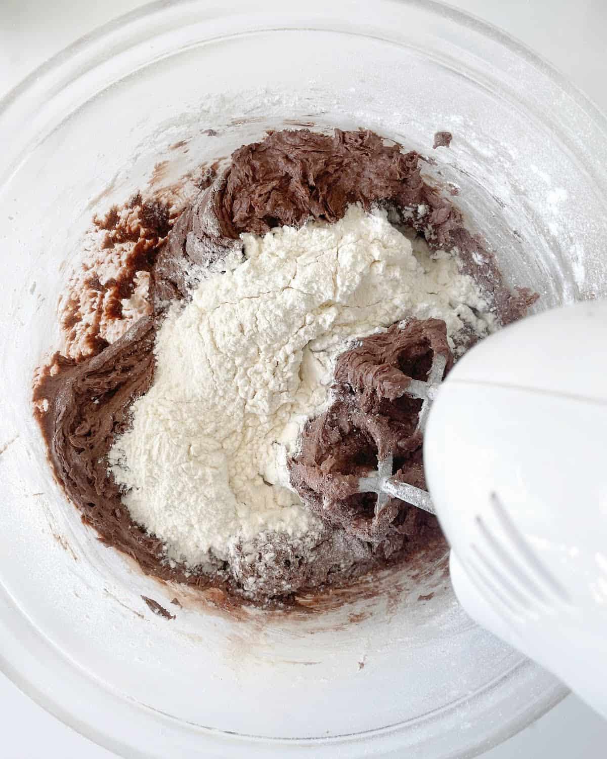 Flour added to chocolate cookie mixture in a glass bowl with an electric mixer. White marble surface.