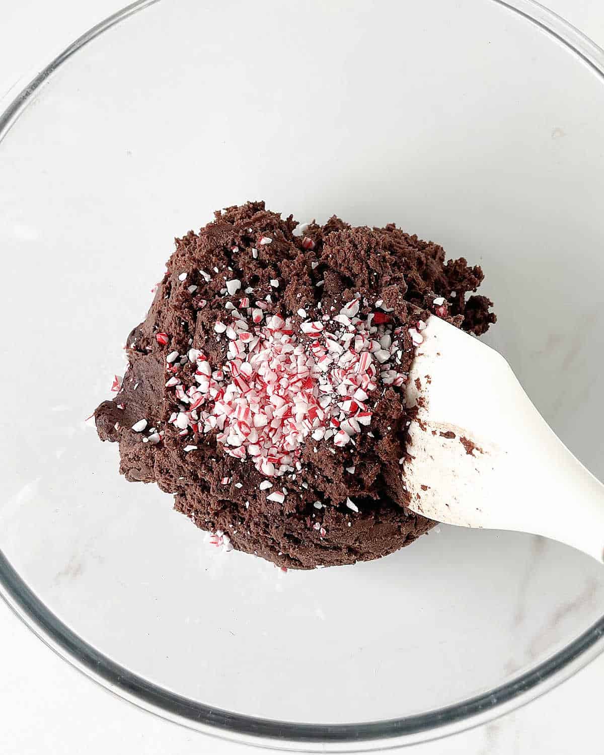 Crushed peppermint candy added to chocolate cookie dough on a glass bowl with a white spatula. White marble surface.