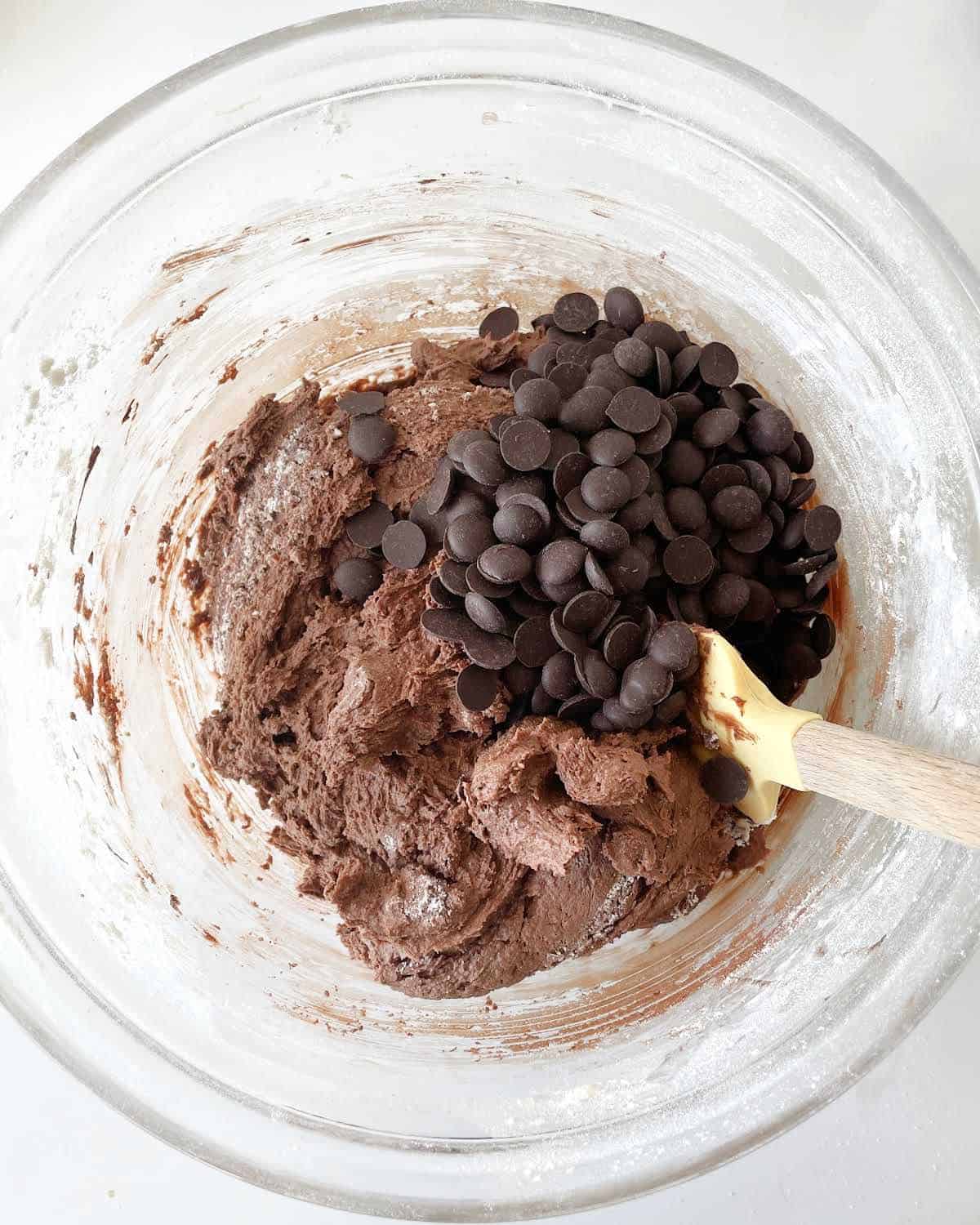Chocolate chips added to chocolate cookie batter in a glass bowl with a spatula. White marble surface.