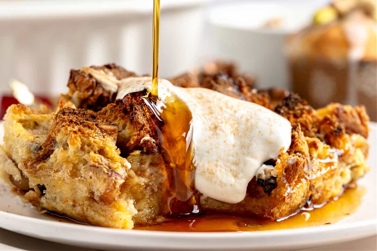 Pouring syrup over cream-topped serving of panettone bread pudding. White plate.