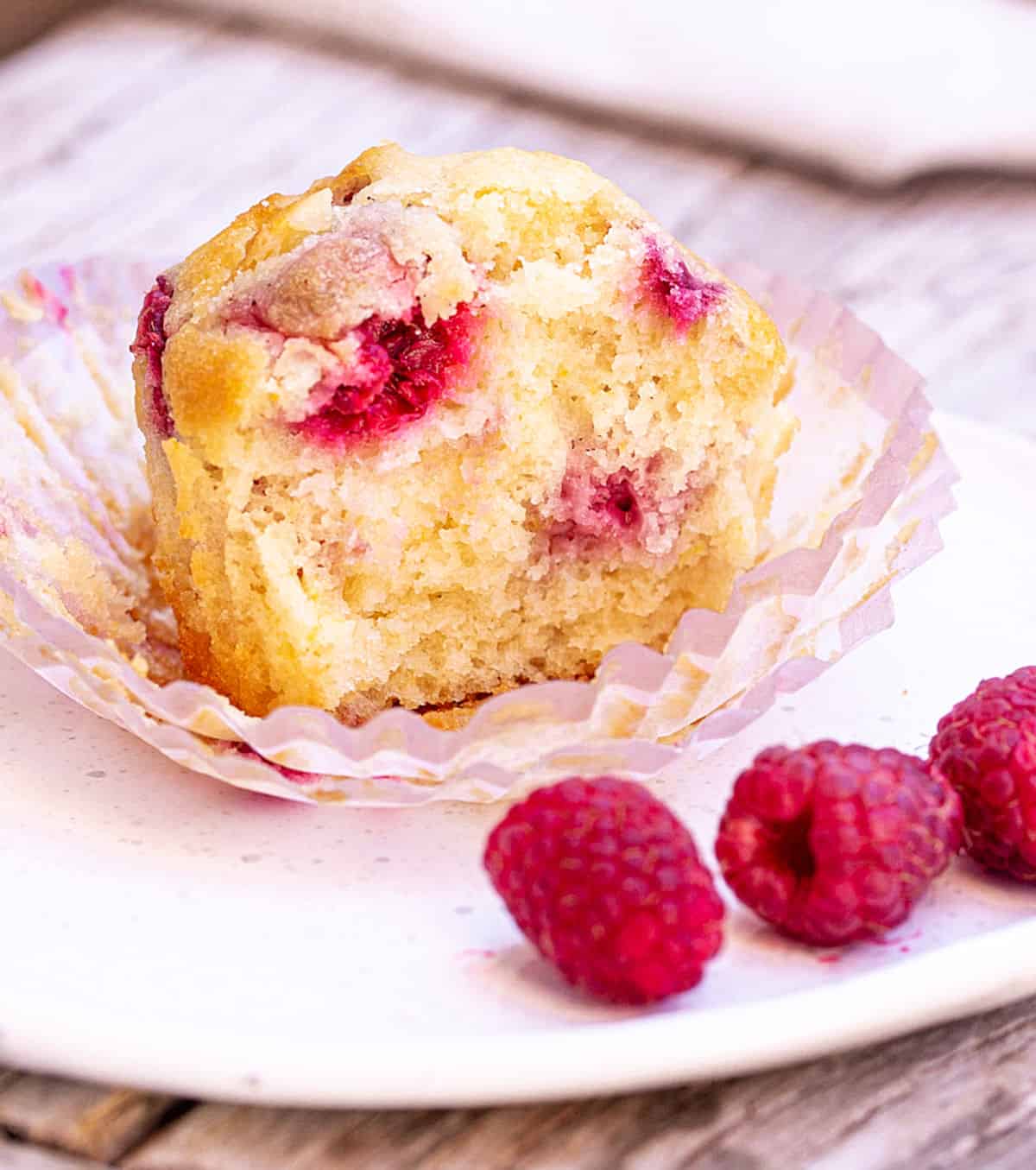 Half raspberry muffin in paper liner on white plate, fresh raspberries around