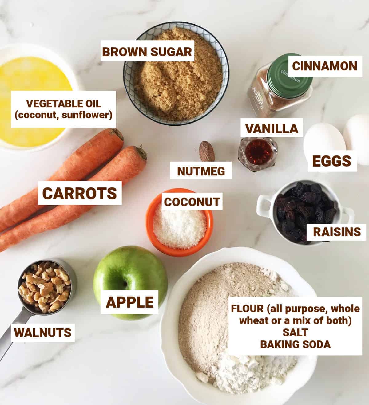 Ingredients in bowls on white marble surface including apple, carrots, oil, spices, walnuts, flour, raisins, sugar