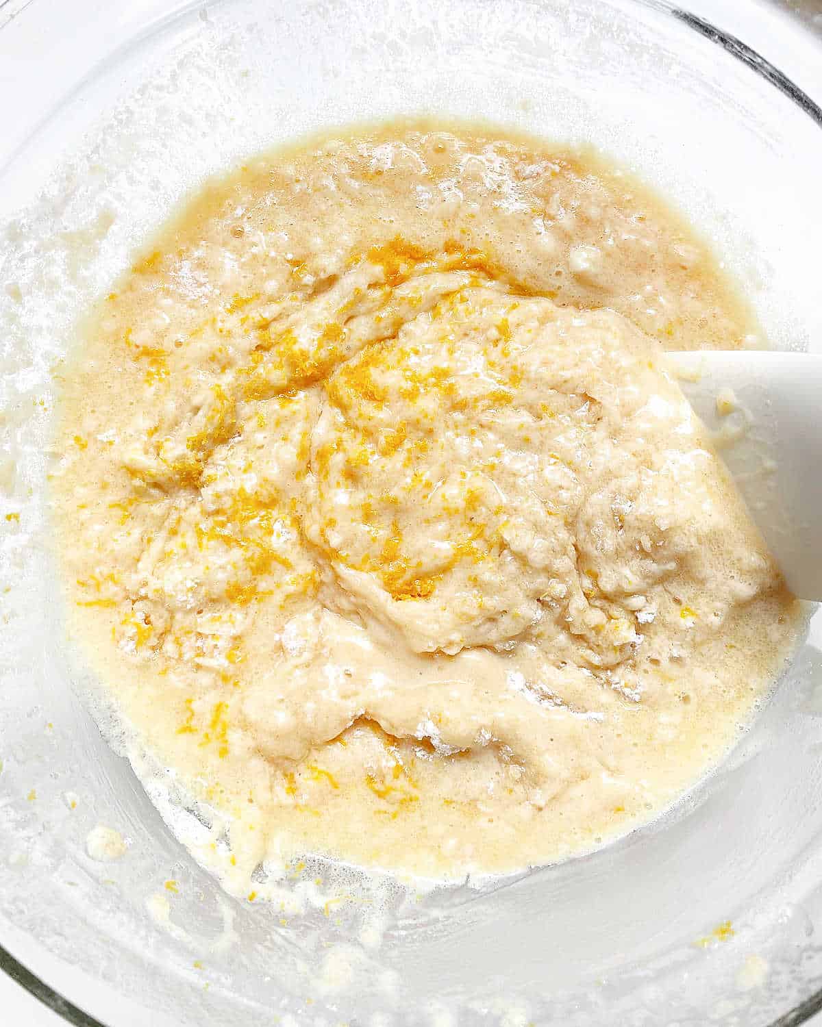 Stirring lemon zest into muffin batter with spatula in a glass bowl.