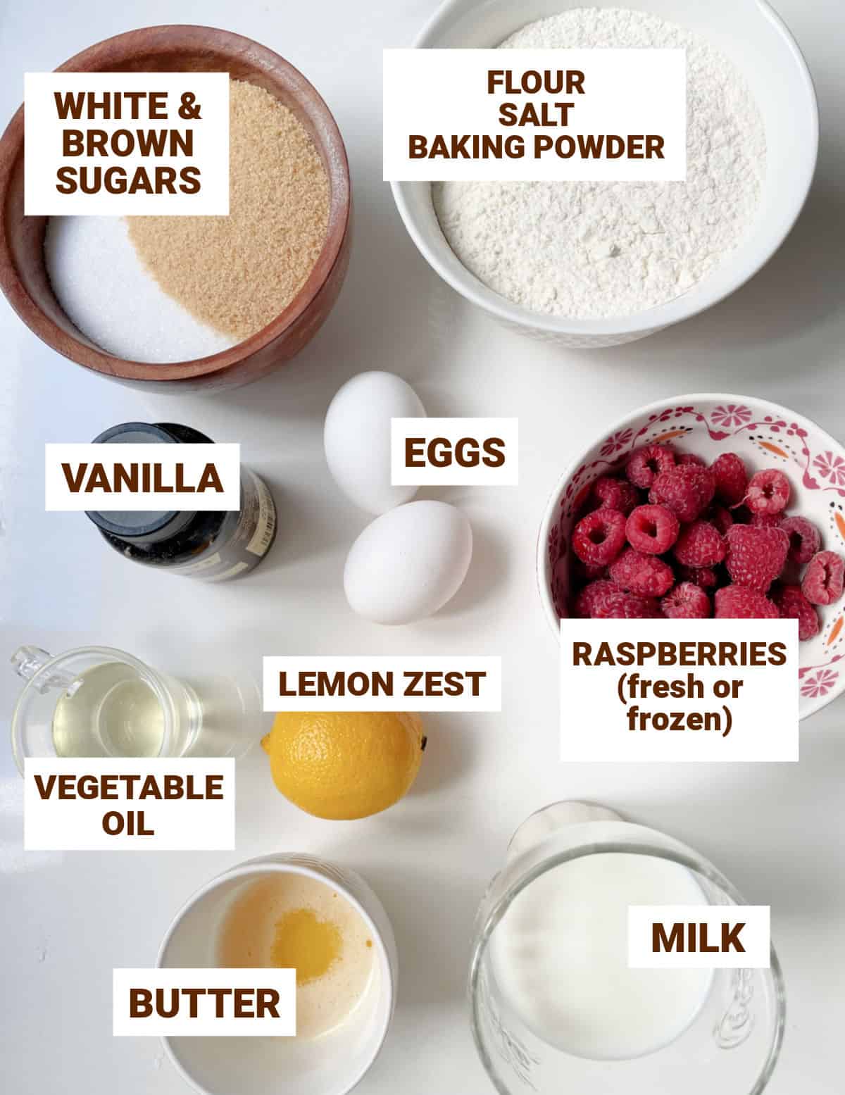 Bowls on white surface with ingredients for lemon raspberry muffins including sugars, butter, oil, milk, eggs, flour mixture