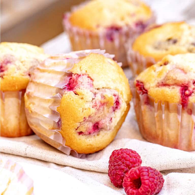 Several raspberry muffins in paper liners on a beige cloth. Fresh raspberries.