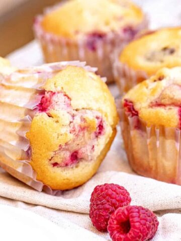 Several raspberry muffins in paper liners on a beige cloth. Fresh raspberries.