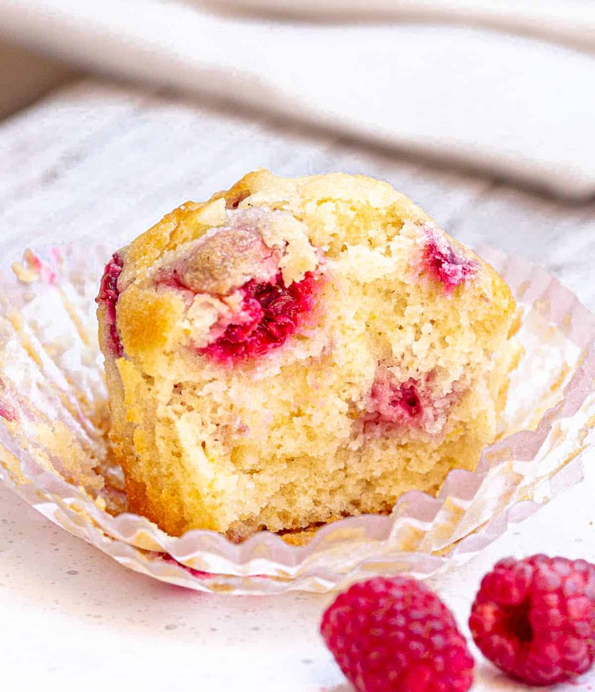 Half raspberry muffin in paper liner on white plate, fresh raspberries around.