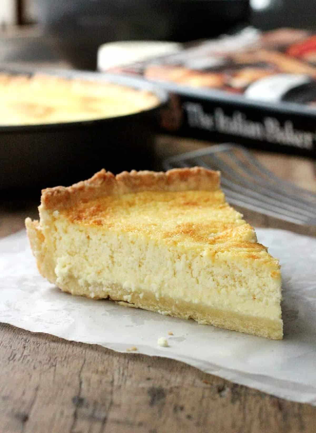 Single slice of ricotta pie on parchment over wooden table, with book, pan in background