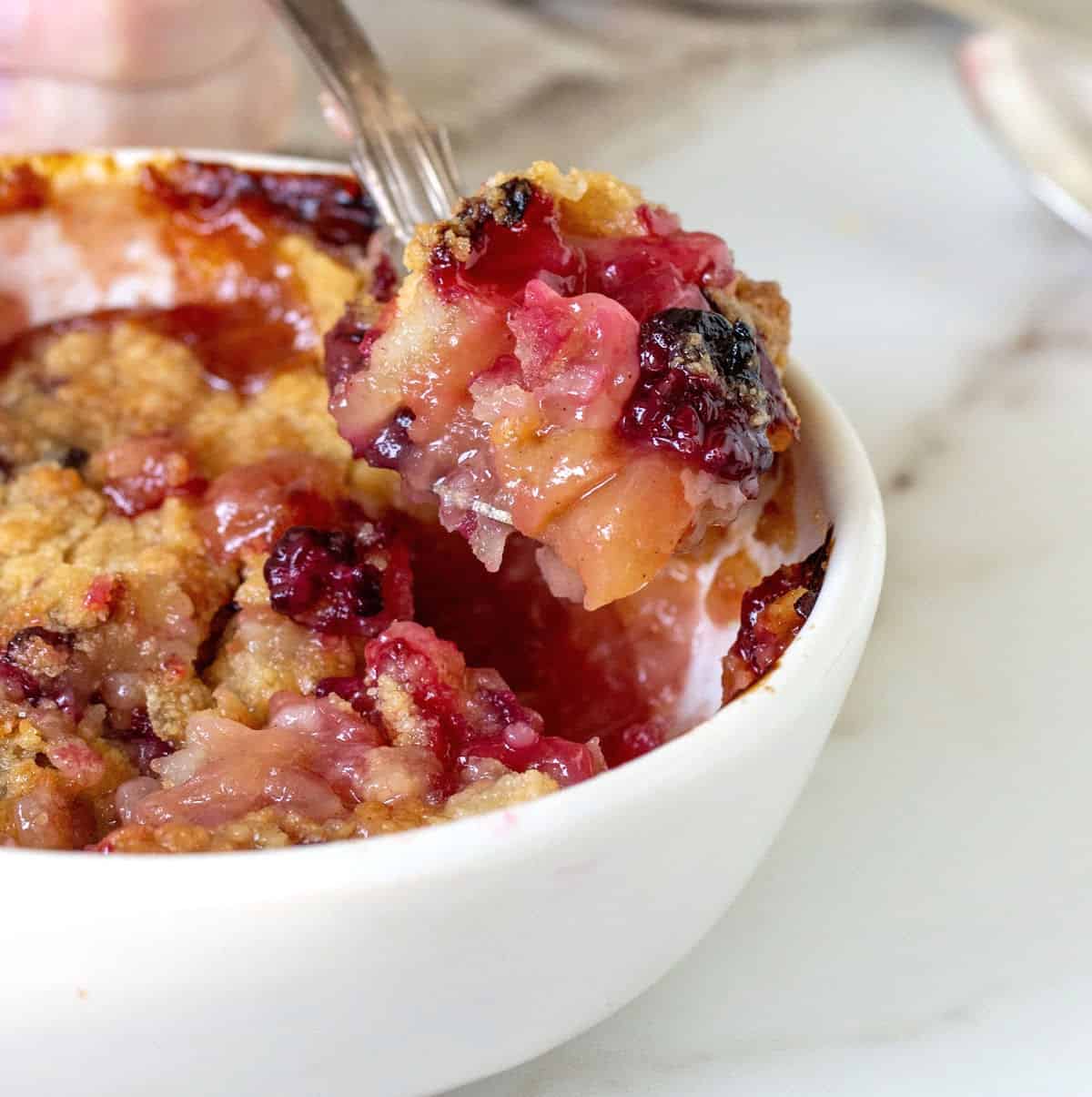 Spoon lifting apple blackberry crumble from white bowl.