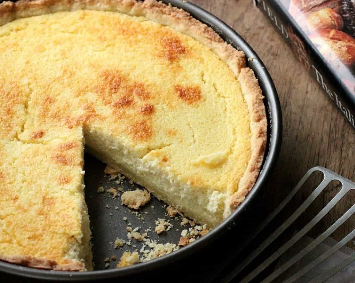 Pie plate with ricotta pie on a wooden table, one slice missing, a metal spatula