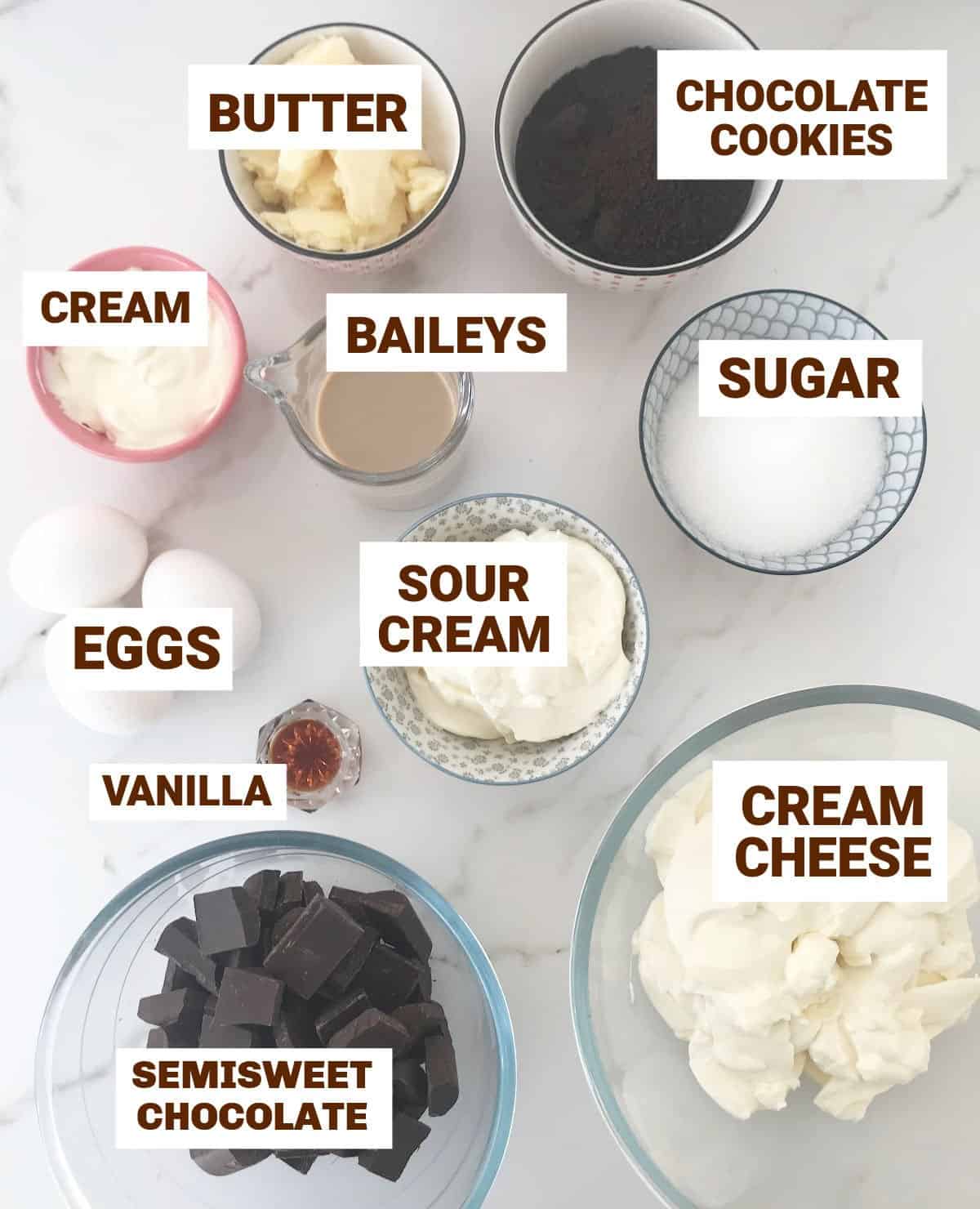 Ingredients for baileys chocolate cheesecake in bowls on white surface, including eggs, cookies, butter, sugar, vanilla, sour cream, sugar, cream.