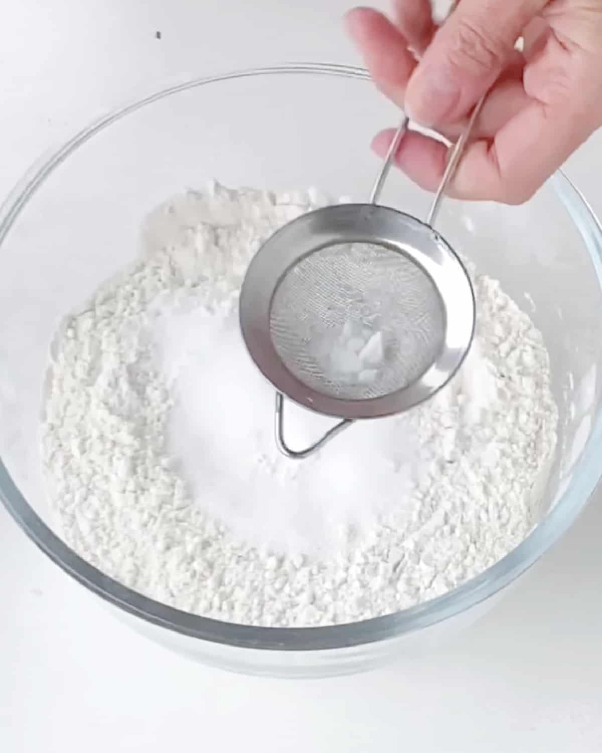 Sifting baking soda over flour mixture in a glass bowl. White surface.