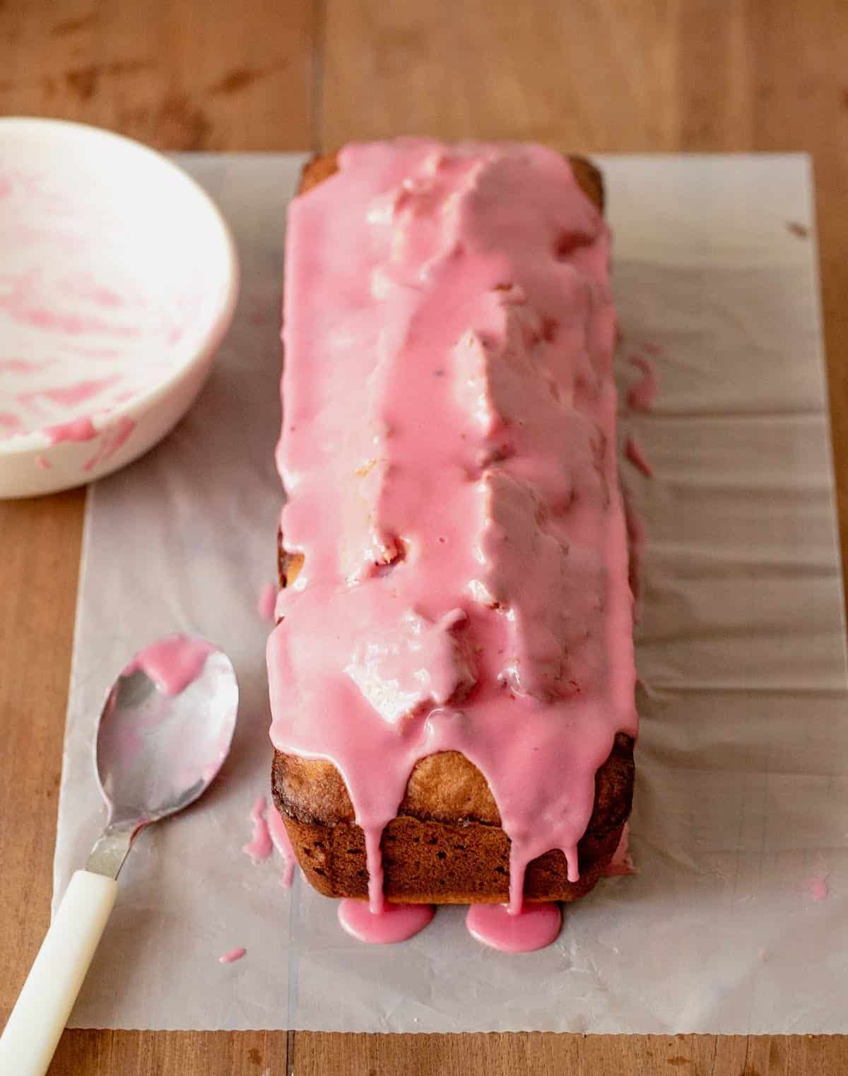 Pink glazed loaf on parchment paper on wooden table, white bowl and spoon.