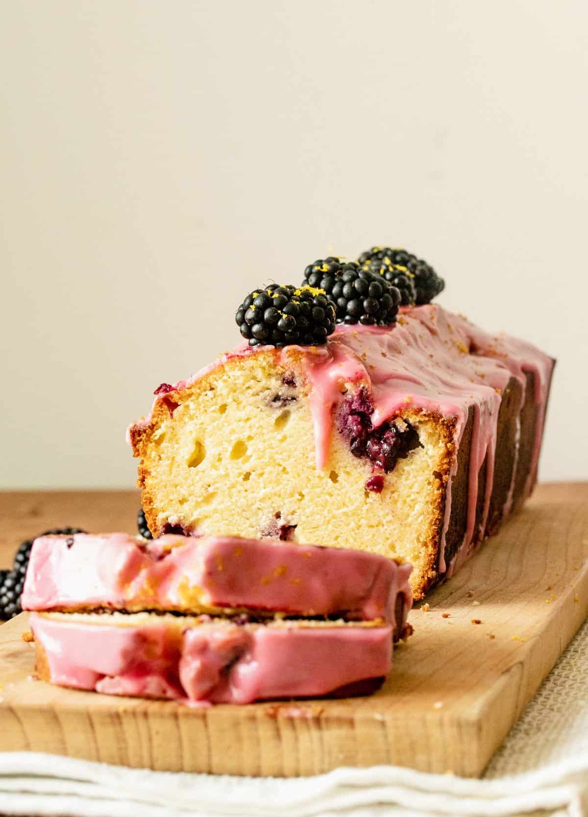 Cut slices and whole blackberry loaf on wooden board, beige background.
