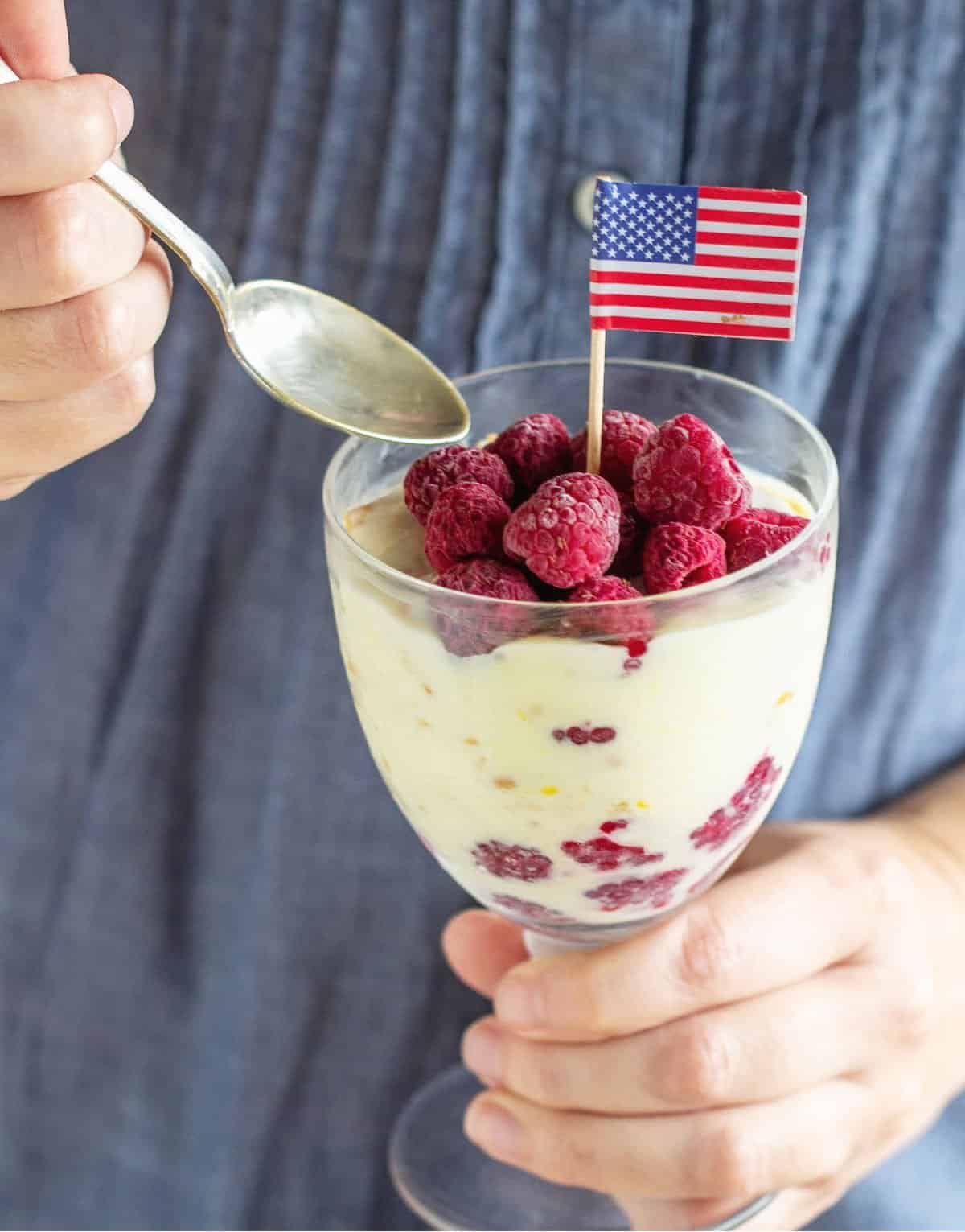 Blue background with hands holding spoon and glass with lemon raspberry dessert, american flag on top.