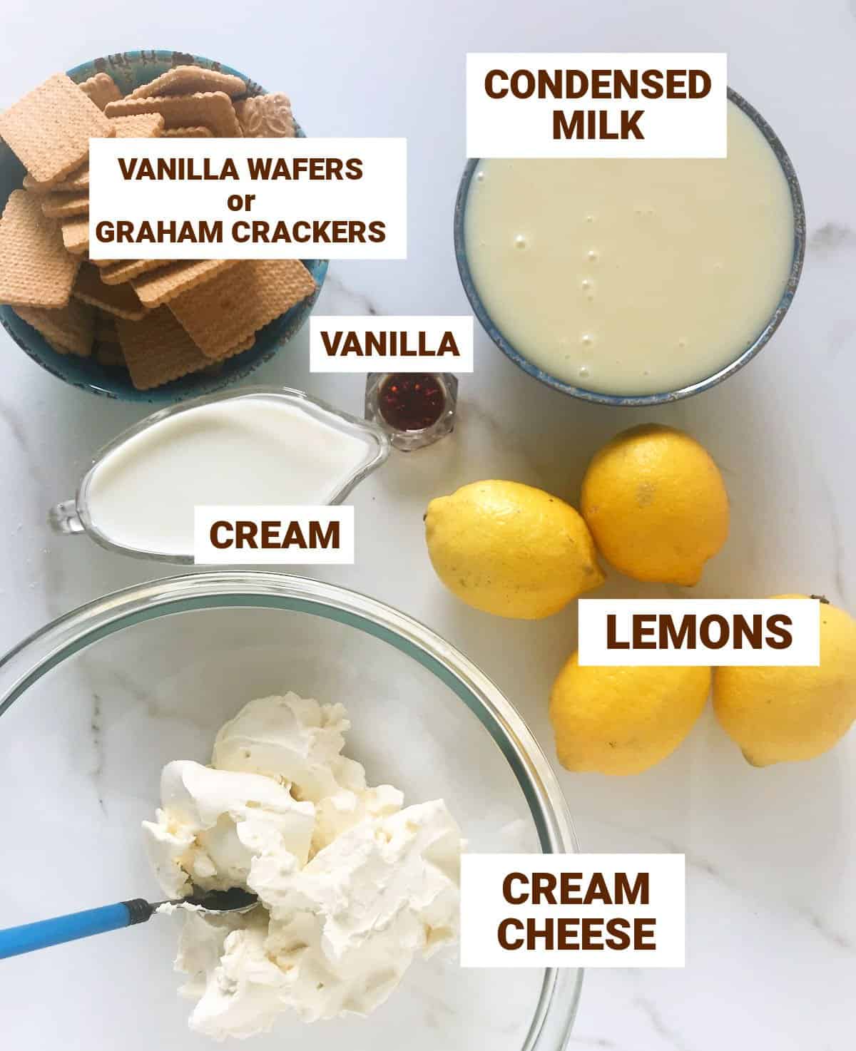 Lemon dessert ingredients in bowls on white surface including condensed milk, cookies, lemons, cream cheese, vanilla, cream.