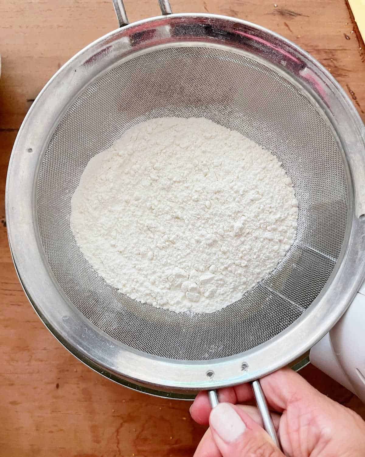 Sifting flour mixture over bowl set on a wooden surface.