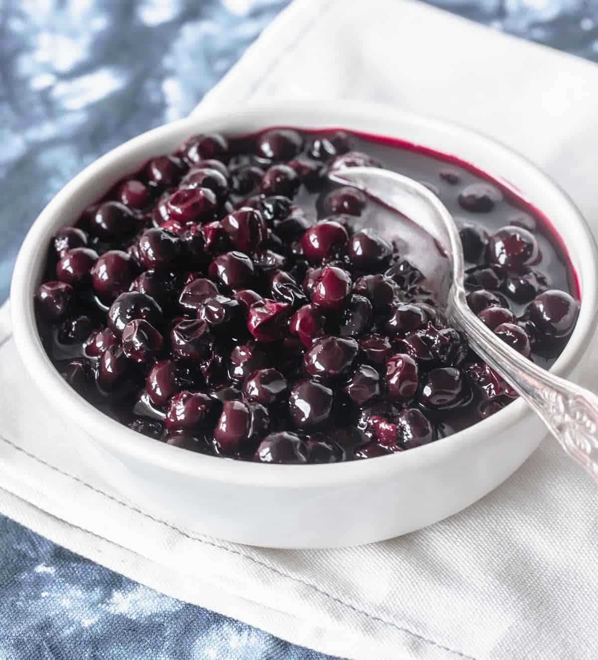 Whole white bowl with blueberry compote and silver spoon on blue white cloth.