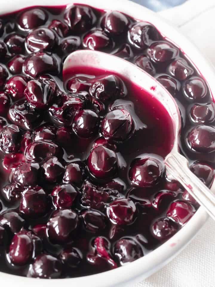 Very close up image of blueberry compote in white bowl with silver spoon.