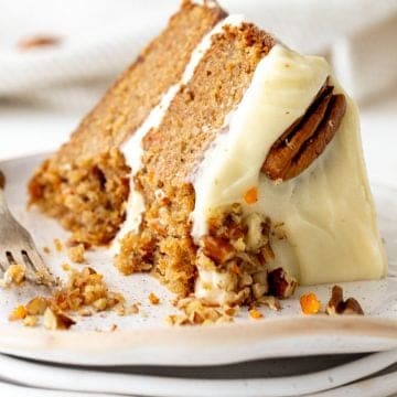 Eaten slice of filled and frosted carrot cake on stack of white plates, pecans and silver fork.