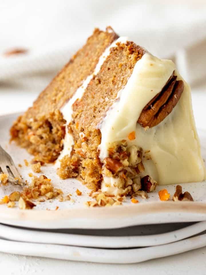 Eaten slice of filled and frosted carrot cake on stack of white plates, pecans and silver fork.