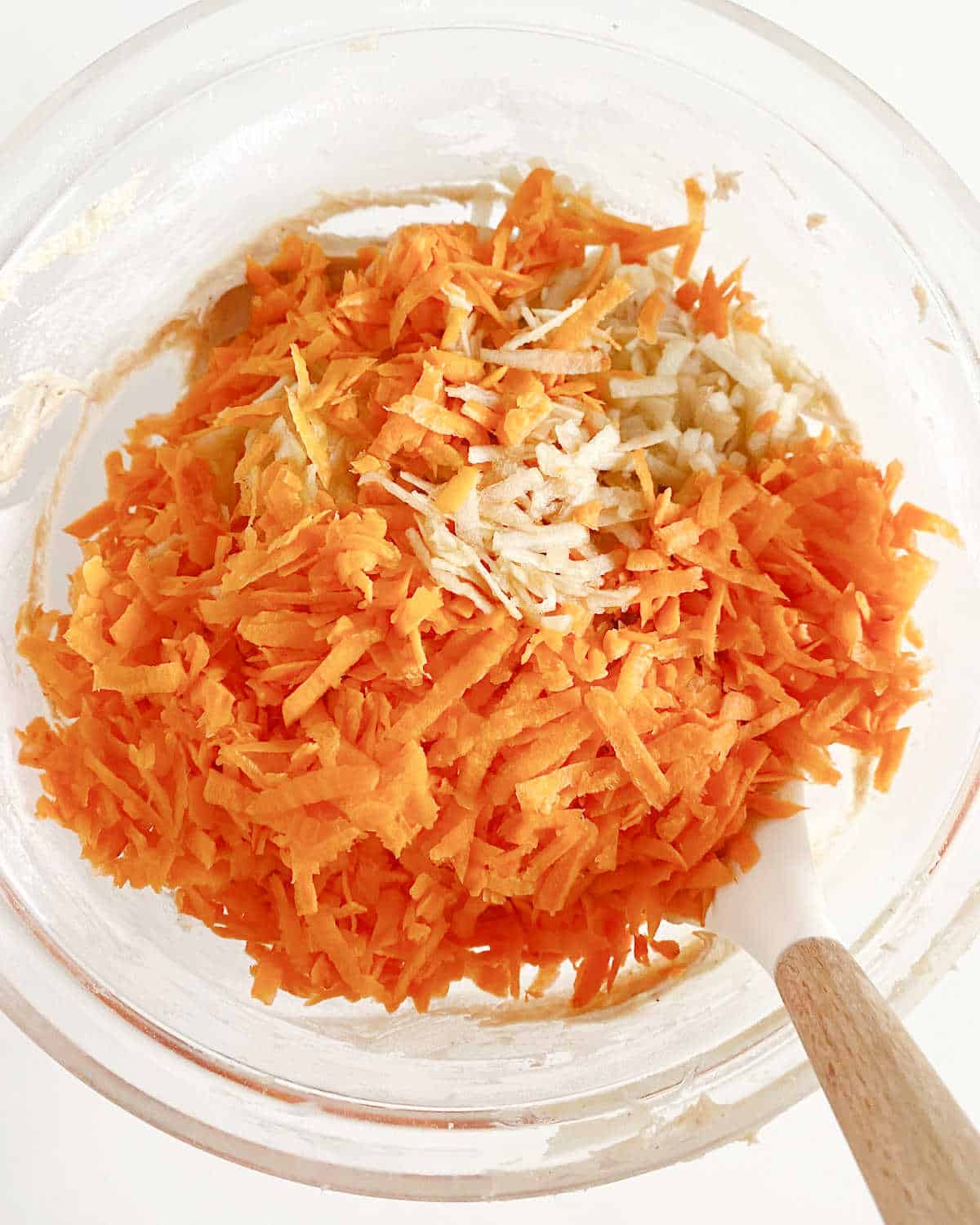 Grated carrot added to cake batter in a glass bowl with a white wooden spatula. White surface.