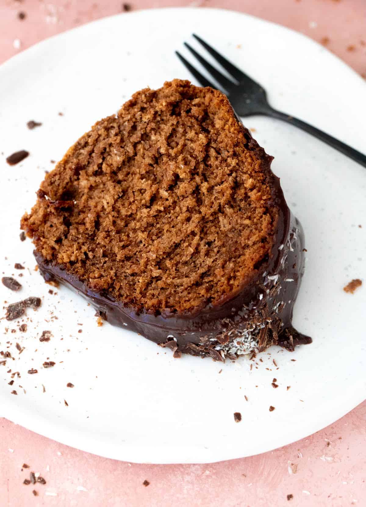 Single slice of chocolate bundt cake on white plate, a dark fork, pink surface.