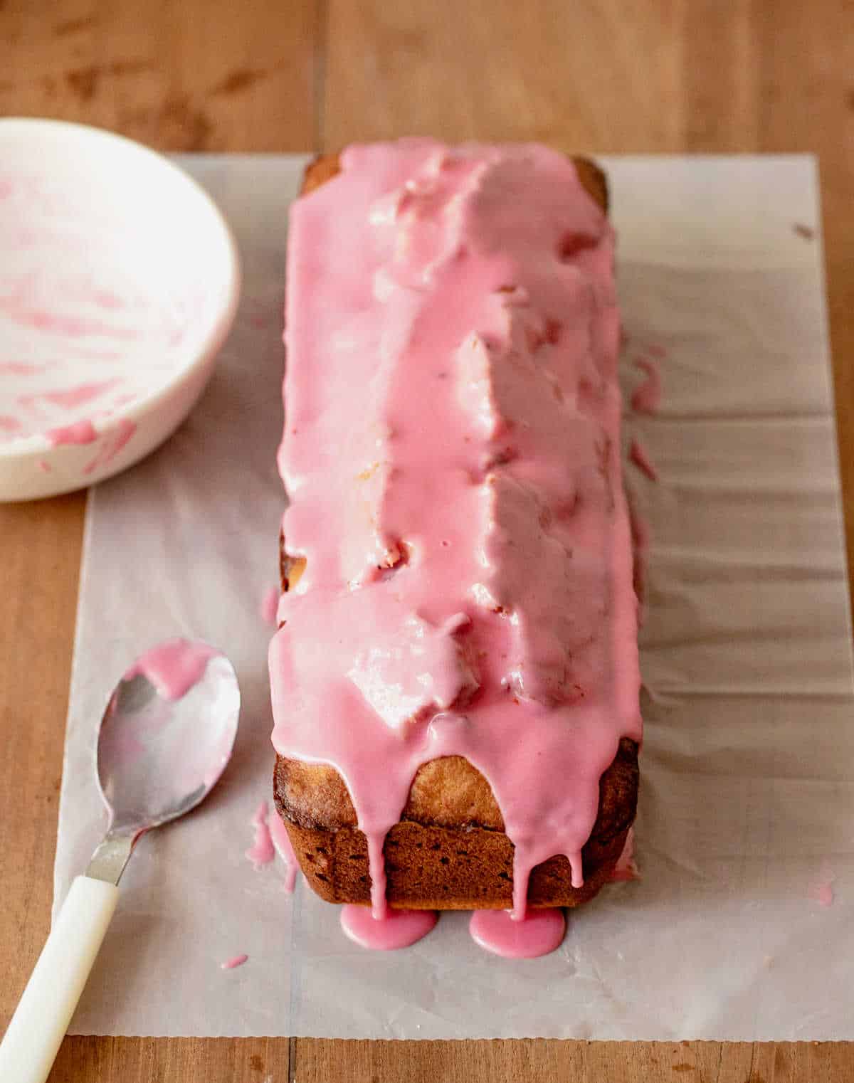 Loaf cake on white parchment paper covered with pink glaze. A spoon and white bowl. 