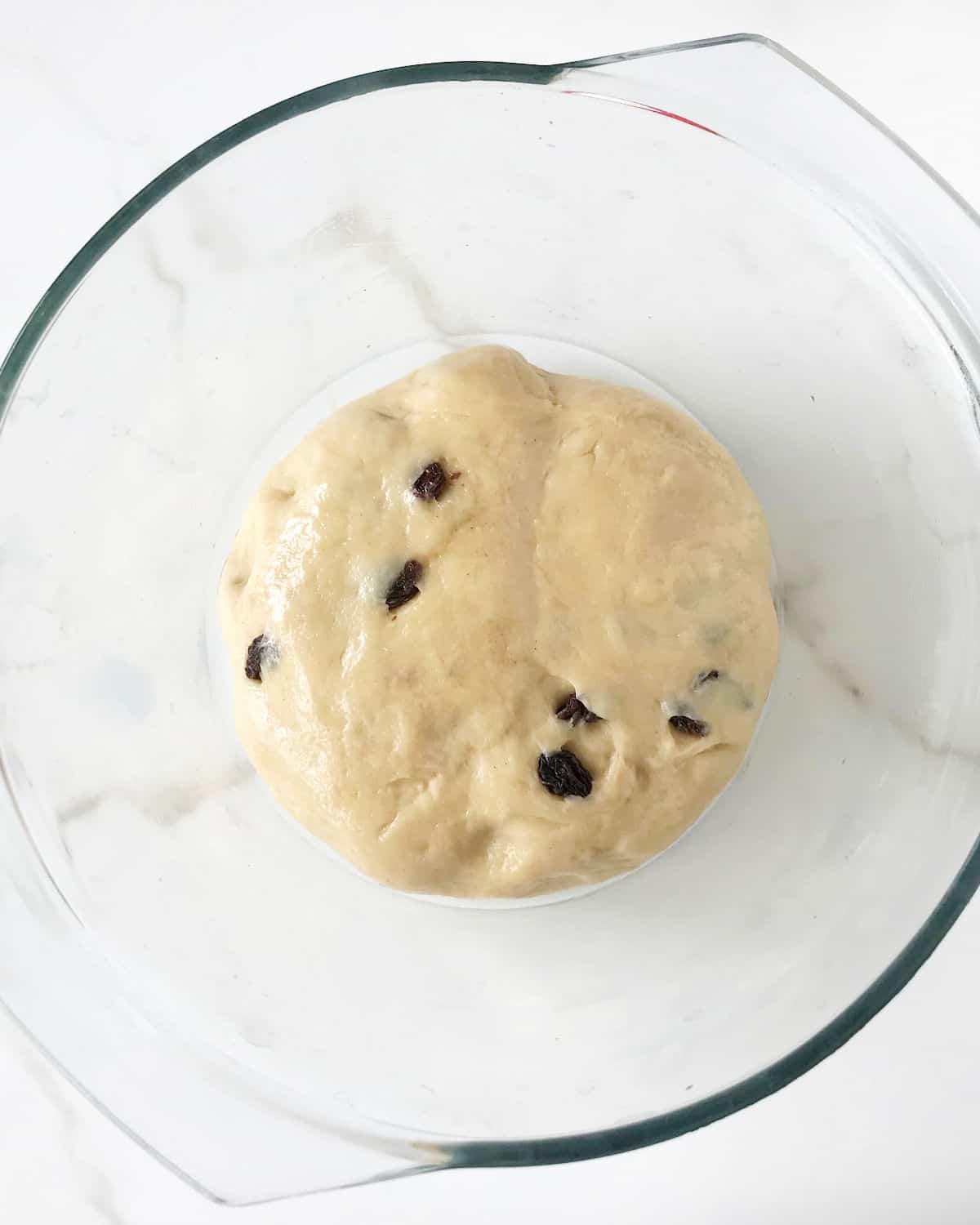 Round of hot cross buns with raisins in a glass bowl set on white marble.