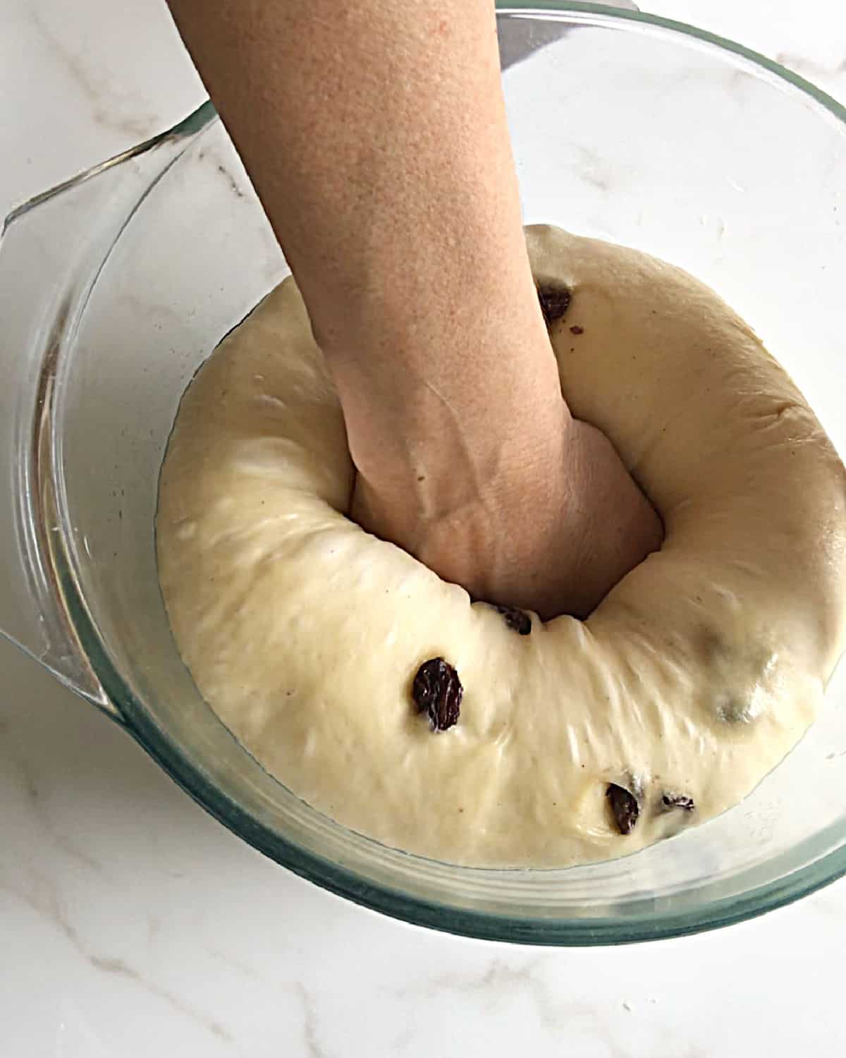 Punching down a risen raisin dough in a glass bowl on a white marble surface.