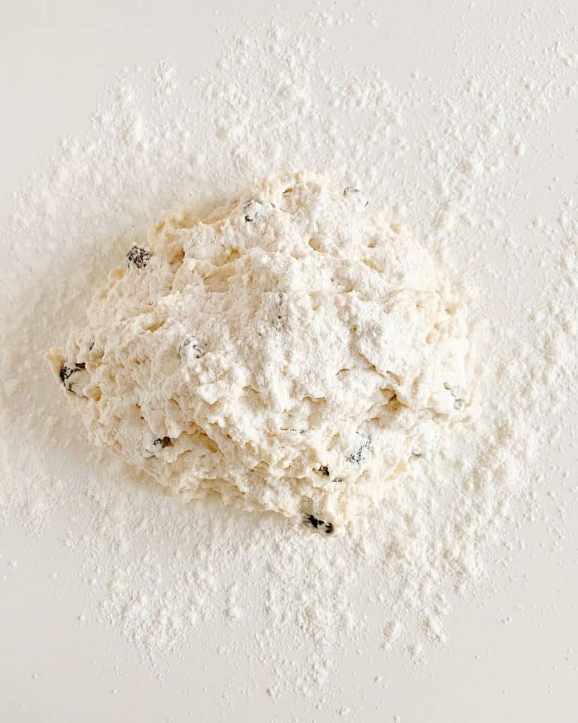 Shaggy mass of raisin soda bread with flour on top on a floured white surface.