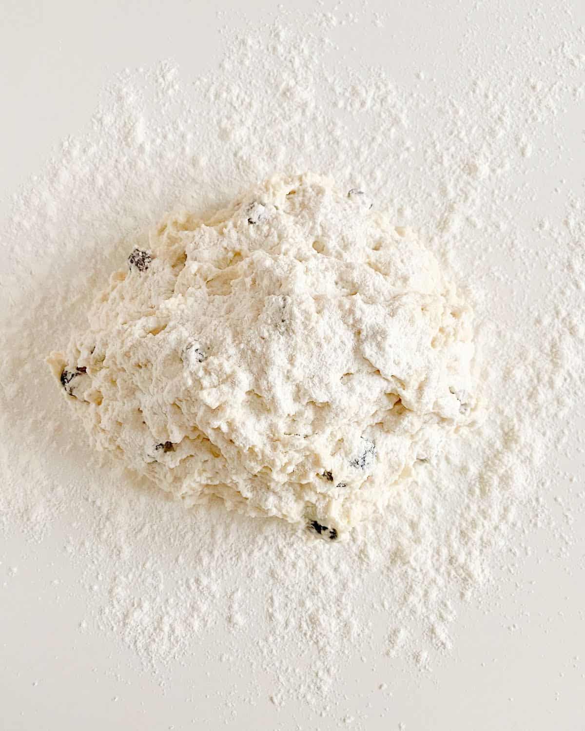 Shaggy mass of raisin soda bread with flour on top on a floured white surface.