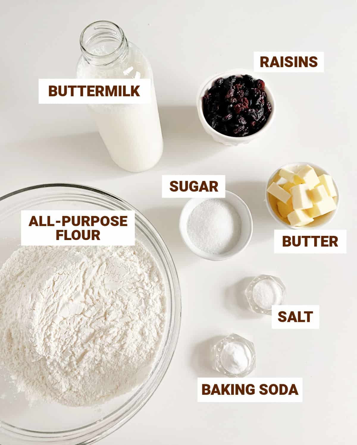 White surface with bowls containing ingredients for Irish soda bread including raisins, buttermilk, butter, sugar, salt, flour, baking soda.