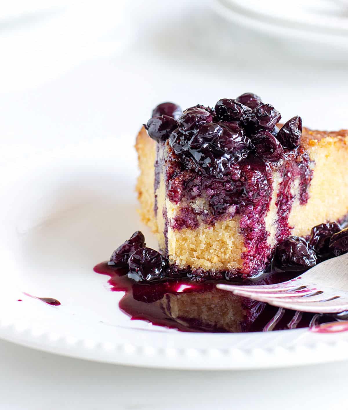 Eaten slice of polenta cake, a pool of blueberry sauce, white plate and background, silver fork.