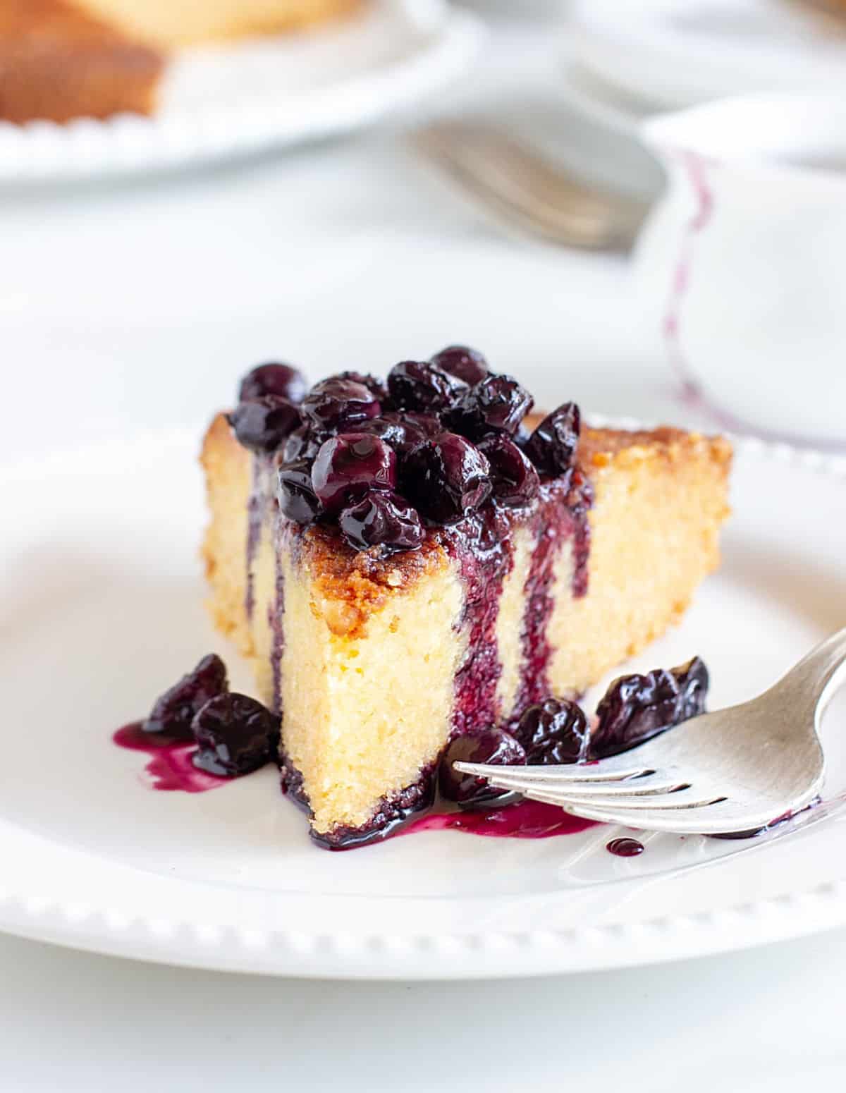 Slice of lemon cake with blueberry sauce on white plate, silver fork, white props on background.