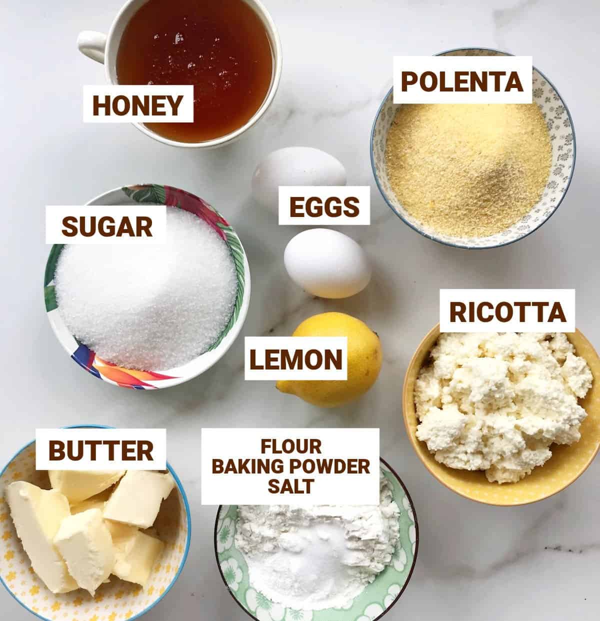 Lemon polenta cake ingredients in bowls on white surface including ricotta, butter, honey, eggs, sugar, flour.