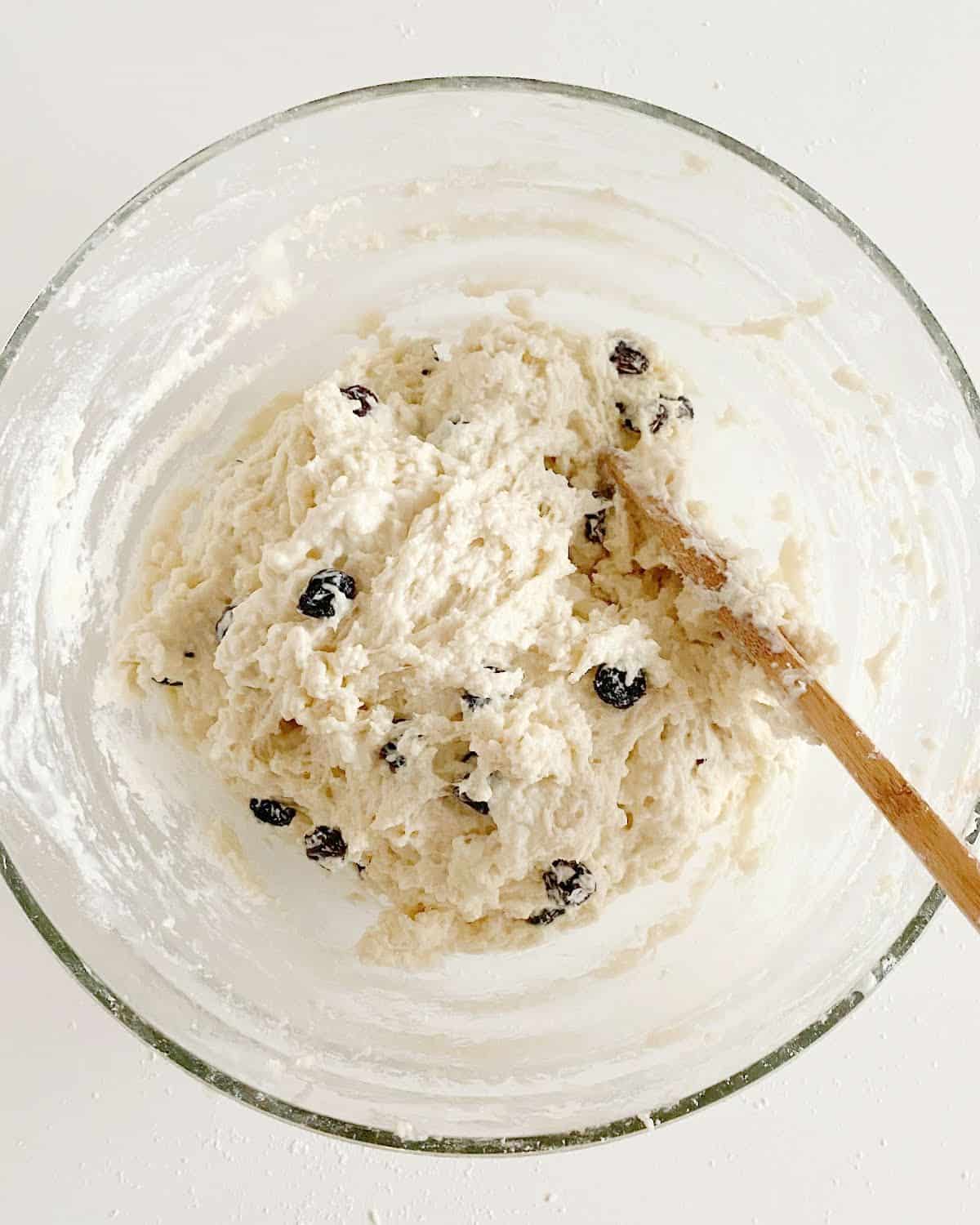 Raisin soda bread dough in a glass bowl with a wooden spoon. White surface.